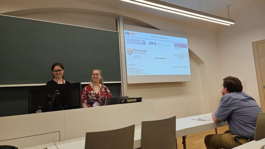 Photo of Carina and Anne at the presenter's desk smiling at the audience. The screen shows the last slide of their presentation where they express thanks to different entities.