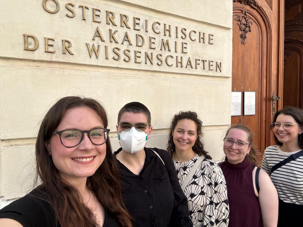 Selfie of all five of us smiling at the camera in front of the writing "Österreichische Akademie der Wissenschaften" on a large building of the university.
